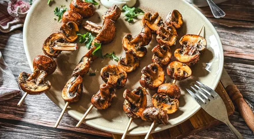 Gegrillte Champignons auf Spießen, mit Kräutern verfeinert und auf einem Teller mit Dip serviert. Ein Genuss für jeden Pilzliebhaber!