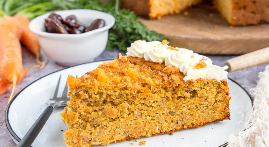 Ein saftiges Stück karottenkuchen ohne zucker, verziert mit cremiger Glasur und Orangenschale. Daneben liegen frische Karotten und Datteln.