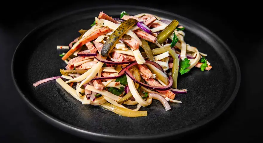 Ein appetitlicher Schweizer Wurstsalat mit Wurststreifen, Käse, roten Zwiebeln und Gewürzgurken auf einem dunklen Teller.