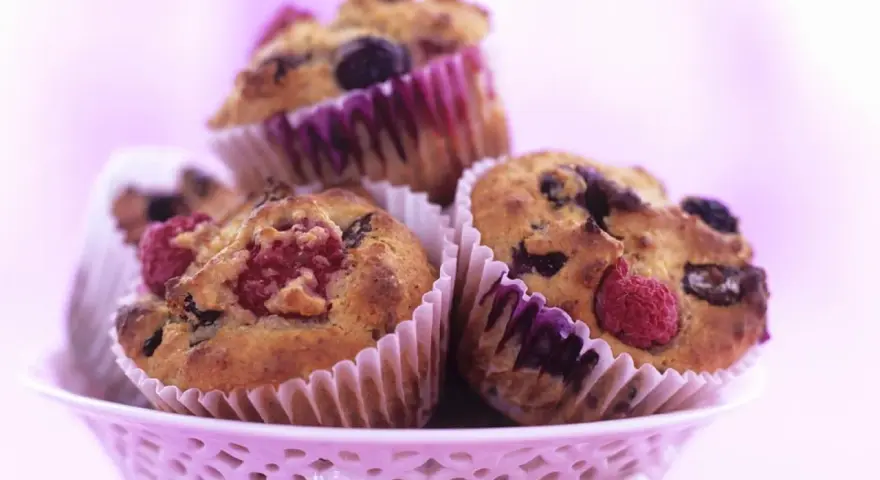 Saftige Muffins mit Obst und Joghurt, garniert mit frischen Himbeeren und Blaubeeren, in einem rosa Körbchen.