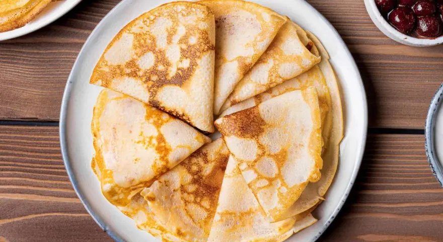 Omas Pfannkuchen-Rezept: goldbraune, gefaltete Pfannkuchen auf einem Teller, daneben eine Schüssel mit Kirschen.