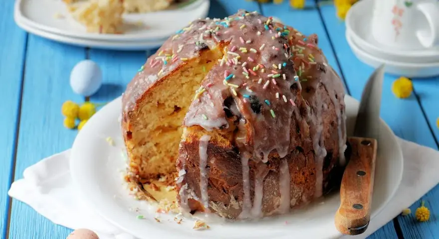 Ein Anschnitt eines Paska-Rezepts mit Zuckerguss und bunten Streuseln. Daneben liegen bunte Ostereier und gelbe Blümchen.