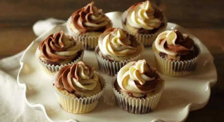 Sieben Marmormuffins mit zweifarbiger Schokoladen-Vanille-Glasur auf einer weißen Tortenplatte. Ein köstliches Marmormuffins Rezept.