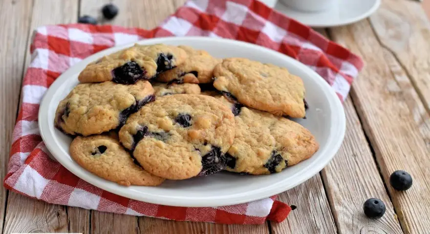 Saftige Blaubeer Cookies auf einem Teller, daneben frische Blaubeeren und eine rot-weiße karierte Serviette.