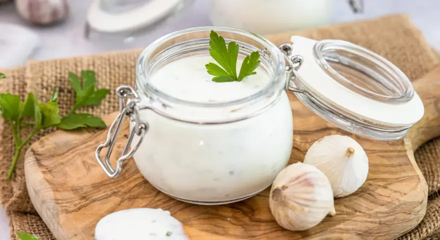 Ein Glas mit cremigem Knoblauch Dip, garniert mit Petersilie, daneben Knoblauchzehen und ein Löffel mit Dip.