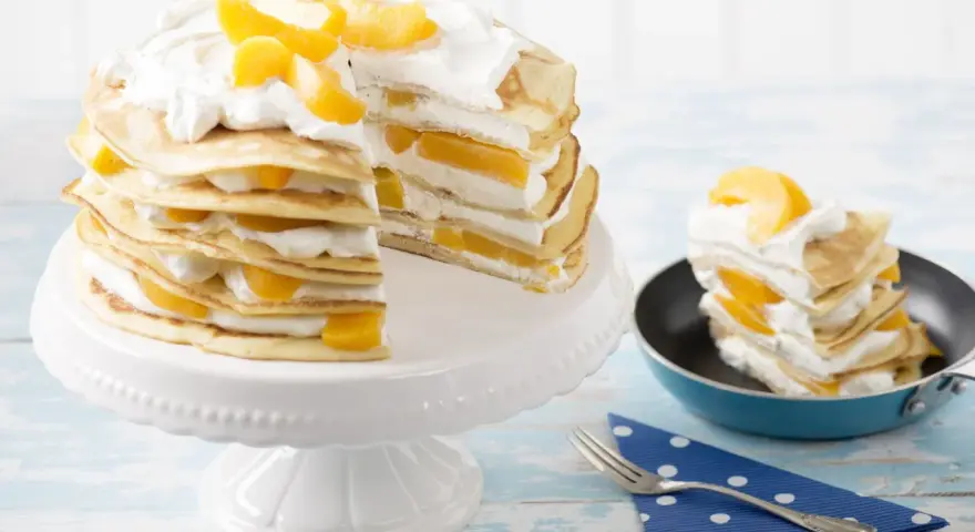 Eine köstliche Pfannkuchen Torte mit Pfirsichen und Sahne, auf einem weißen Tortenständer und ein Stück in einer kleinen Pfanne.