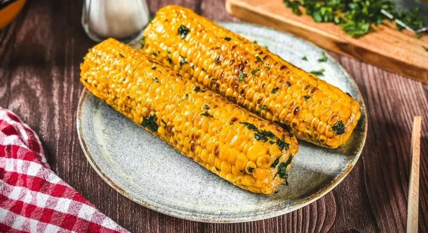 Zwei goldgelbe maiskolben, perfekt gegrillt und mit Kräutern bestreut, liegen auf einem Teller. Ein Genuss für alle, die maiskolben grillen lieben.