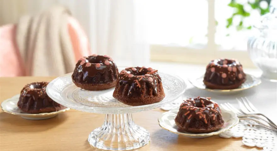 Schokoladen-Mini-Gugelhupf dekorieren mit Schokoglasur und bunten Streuseln auf einem Tortenständer.
