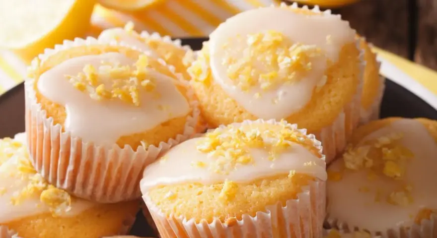 Leckere zitronenmuffins rezept: goldgelbe Muffins mit weißer Glasur und Zitronenschale bestreut, daneben frische Zitronenscheiben.