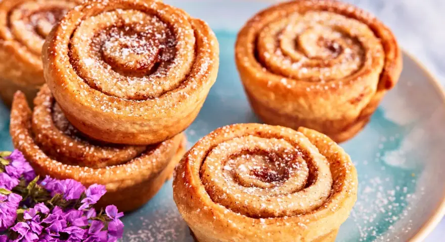 Saftige Zimtschnecken Muffins, bestäubt mit Puderzucker, liegen auf einem blauen Teller. Lila Blumen schmücken die süße Leckerei.