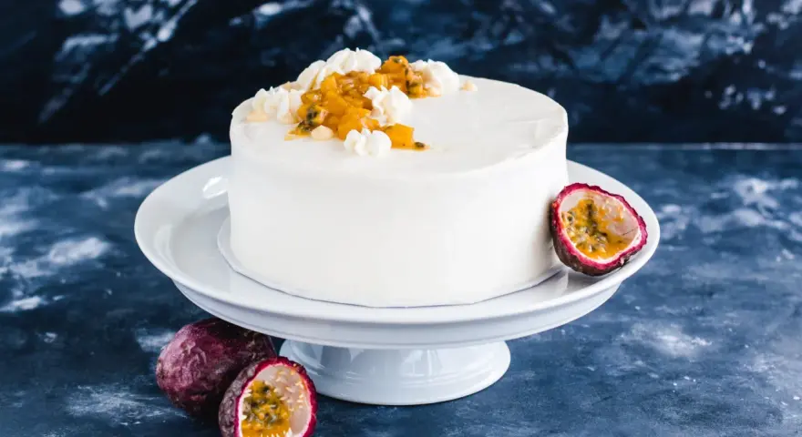 Eine weiße Maracuja Torte mit frischer Maracuja-Frucht und Sahnetupfern, umgeben von ganzen und halbierten Maracujas.