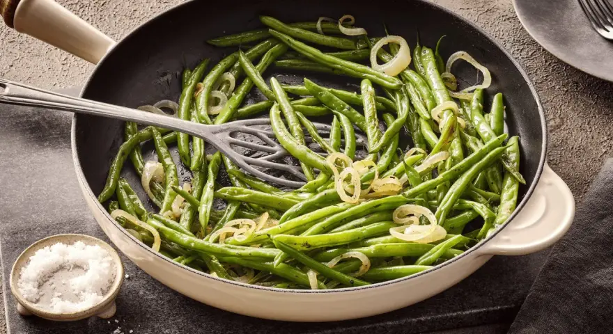 Grüne Bohnen mit Zwiebelringen in einer Pfanne, bereit zum Kochen. Daneben eine Schale mit grobem Salz.