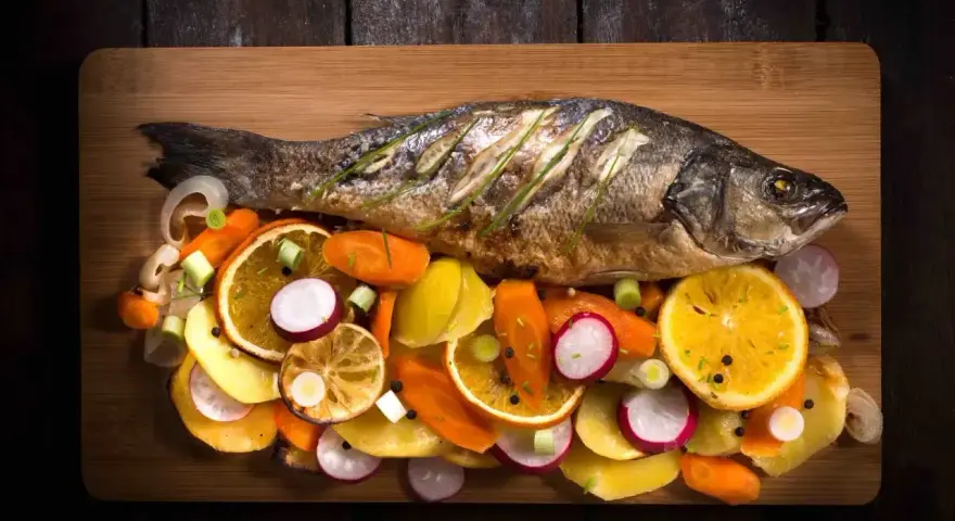 Eine ganze Forelle im Backofen, serviert mit bunten Gemüsescheiben wie Karotten, Radieschen und Orangen auf einem Holzbrett.
