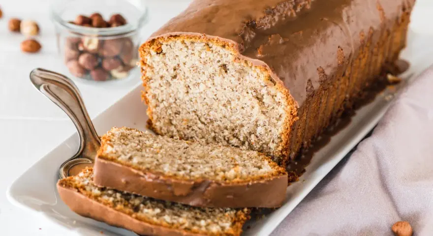 Saftiger Mohn Nuss Kuchen mit Schokoladenglasur, daneben ein Glas mit Haselnüssen und zwei angeschnittene Scheiben.