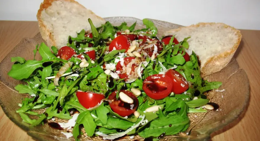 Frischer Rucola Salat mit Kirschtomaten, Pinienkernen und Parmesan, beträufelt mit Balsamico-Dressing. Dazu knuspriges Brot.