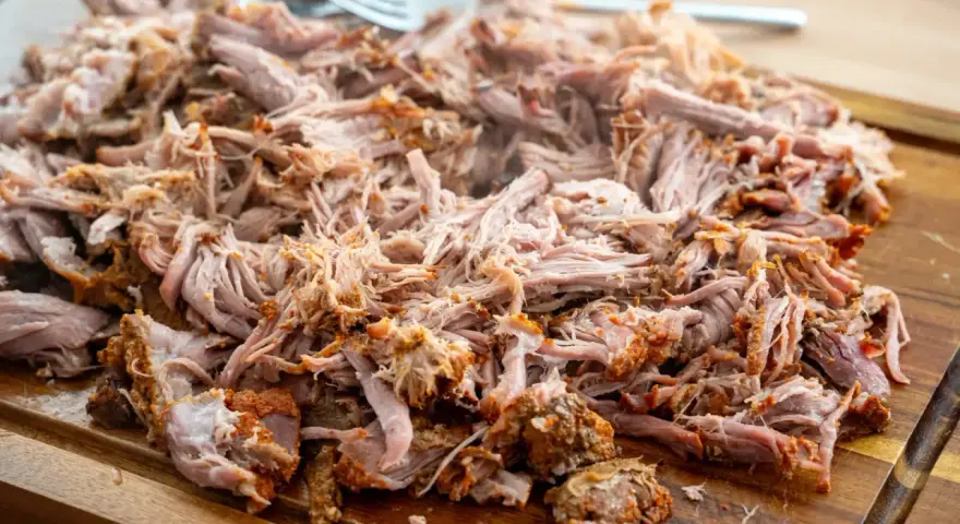 Ein Haufen zartes, **gezupftes Fleisch** liegt auf einem Holzbrett, bereit für leckere Sandwiches oder Tacos. Eine Gabel liegt daneben.