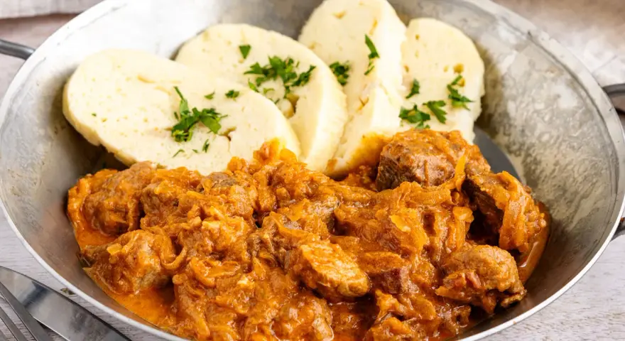 Herzhaftes Szegediner Gulasch mit Klößen, garniert mit Petersilie, serviert in einer Metallschüssel.