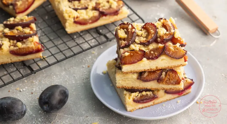 Saftiger zwetschgenkuchen vegan mit Streuseln auf einem Teller gestapelt. Daneben frische Zwetschgen und ein Kuchenblech.