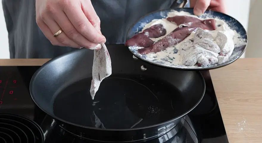 Ein Stück Leber wird in eine heiße Pfanne mit Öl gelegt, um sie zu braten. Auf einem Teller liegen weitere Leberstücke, die in Mehl gewendet wurden.