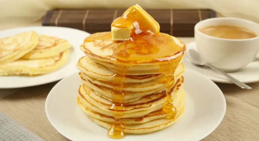 Ein Stapel goldbrauner Pancakes, übergossen mit Honig und Butter, daneben eine Tasse Kaffee. Perfekt für ein leckeres Pancakes Rezept.