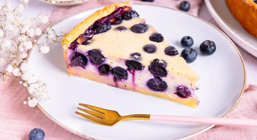 Ein Stück Heidelbeerkuchen Landfrauenart mit saftigen Blaubeeren und cremigem Guss auf einem Teller, daneben eine goldene Gabel und frische Beeren.