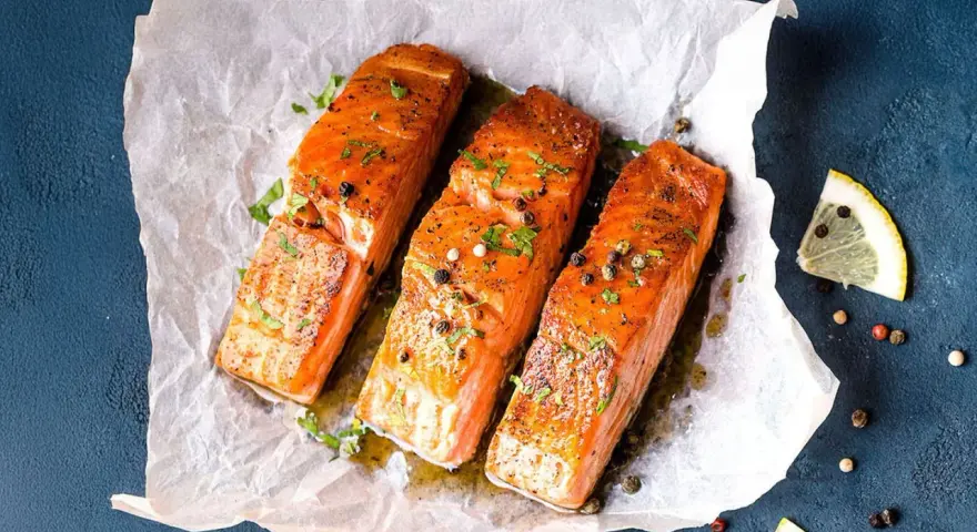 Drei saftige **Lachs im Ofen**-Portionen, garniert mit Kräutern und Pfeffer, auf Backpapier. Daneben Zitronenspalten.