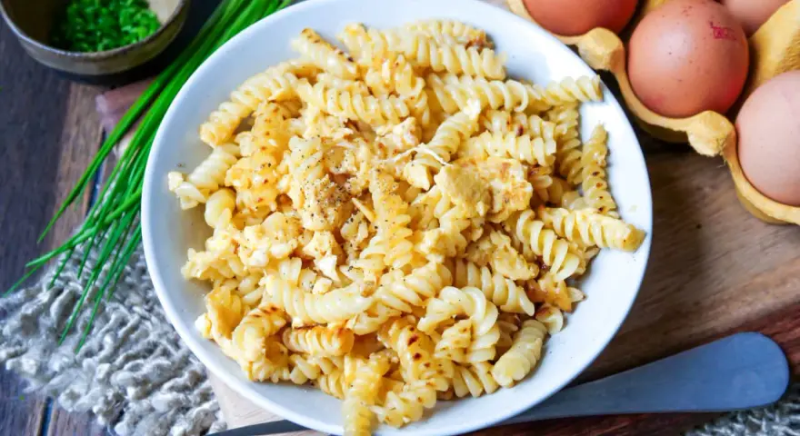 Fusilli-Nudeln mit Rührei, bestreut mit Pfeffer. Daneben liegen frische Eier und Schnittlauch. Ein einfaches Gericht: Nudeln mit Ei.