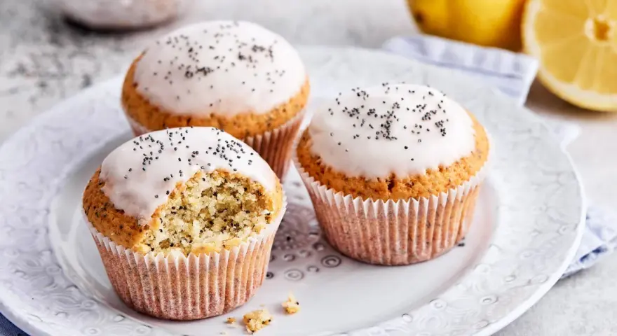 Drei köstliche zitronen mohn muffins mit Zuckerguss und Mohn bestreut, daneben frische Zitronen.