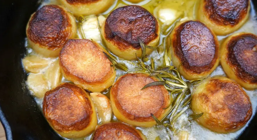 Goldbraun gebratene Fondant-Kartoffeln mit Rosmarin und Knoblauch in einer Pfanne.