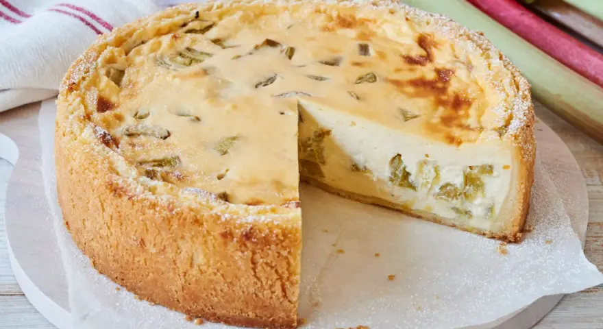 Ein saftiger Rhabarberkuchen, schnell gebacken und mit Puderzucker bestäubt. Daneben liegen frische Rhabarberstangen.