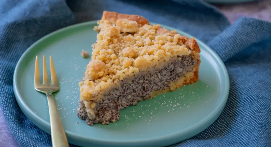 Ein saftiges Stück Mohnkuchen mit Streuseln auf einem mintgrünen Teller, daneben eine goldene Gabel. Ein köstliches Mohnkuchen Rezept.