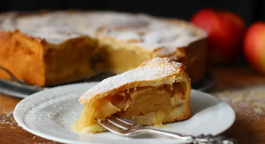 Ein Stück gedeckter Apfelkuchen mit Puderzucker bestäubt, auf einem Teller mit einer Gabel. Im Hintergrund der ganze Kuchen und Äpfel.