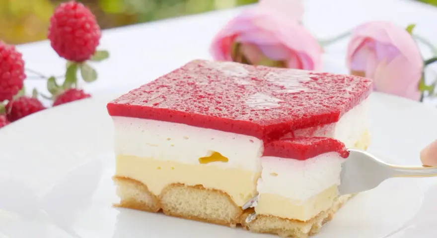 Himbeer-Sahne-Schnitten auf weißer Platte, mit frischen Himbeeren und rosa Rosen im Hintergrund. Ein Stück wird mit einer Gabel herausgenommen.
