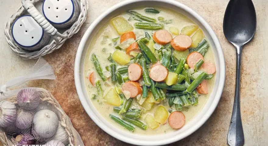 Herzhafte Bohnen-Kartoffel-Suppe mit Würstchen, ein Klassiker unter den Bohnen Rezepten. Dazu Salz- und Pfefferstreuer und frischer Knoblauch.