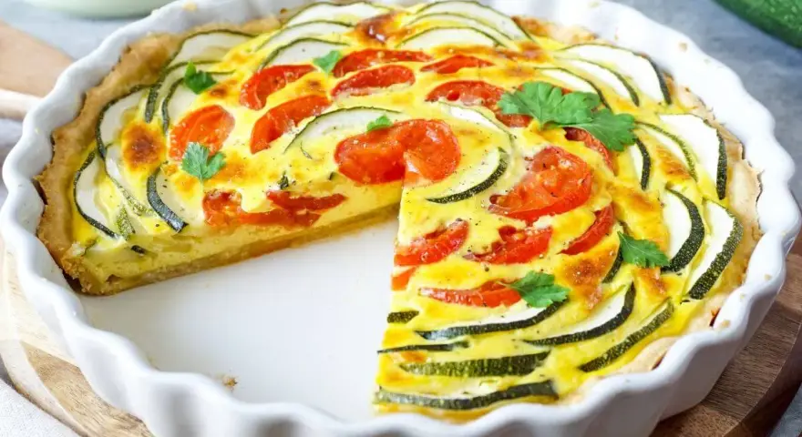 Eine köstliche Zucchini-Quiche mit Tomaten und frischer Petersilie, in einer weißen Quicheform, ein Stück fehlt.