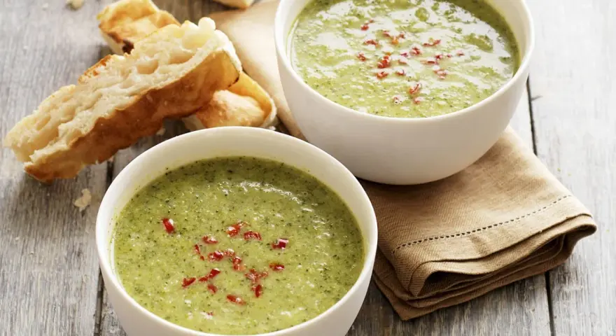 Zwei Schalen cremiger Brokkolisuppe, garniert mit roten Chiliflocken, stehen neben knusprigem Baguette auf einem Holztisch.