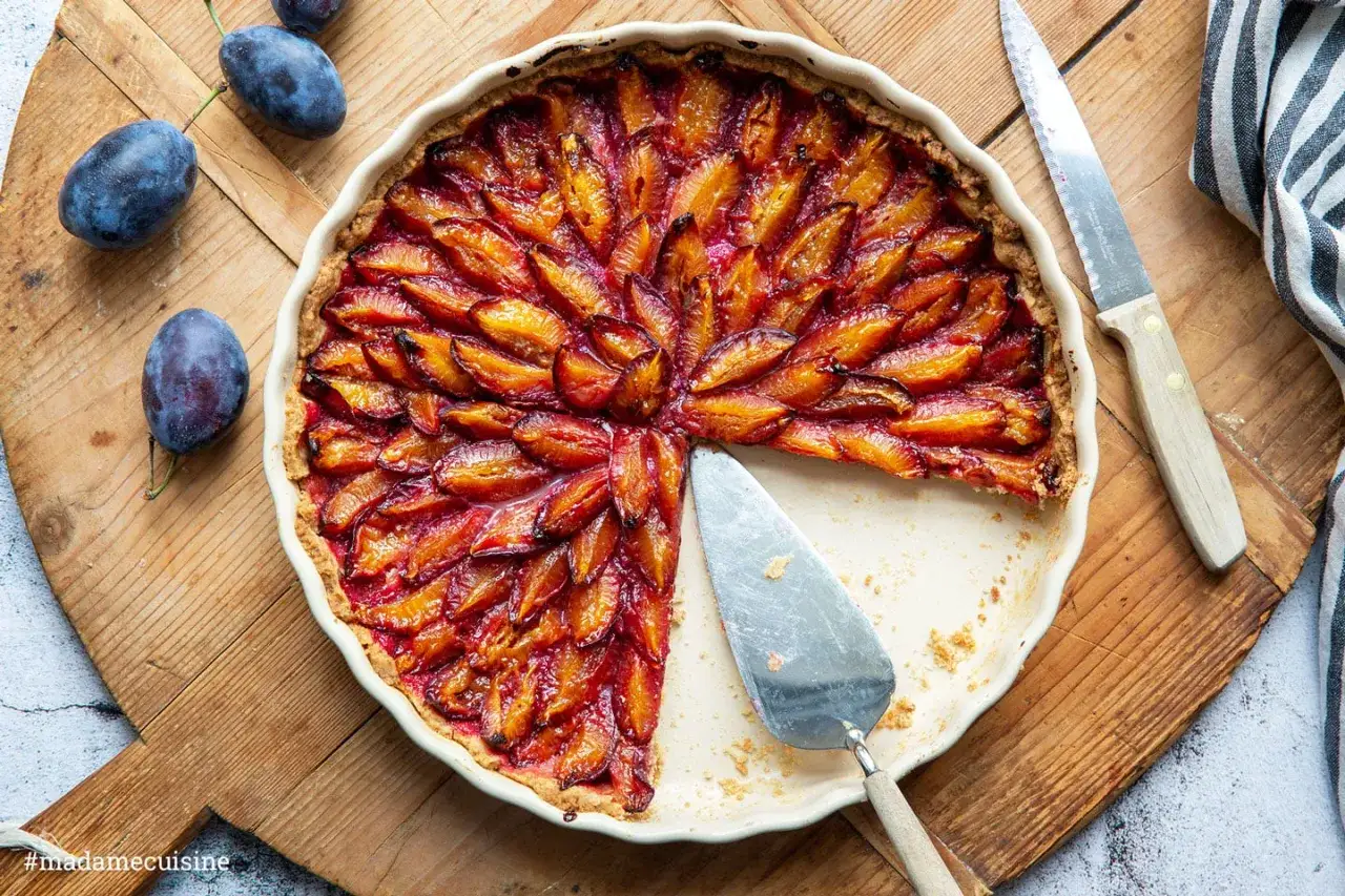Saftige Zwetschgen Tarte, kunstvoll arrangiert, mit einem Stück entnommen. Frische Zwetschgen liegen daneben.