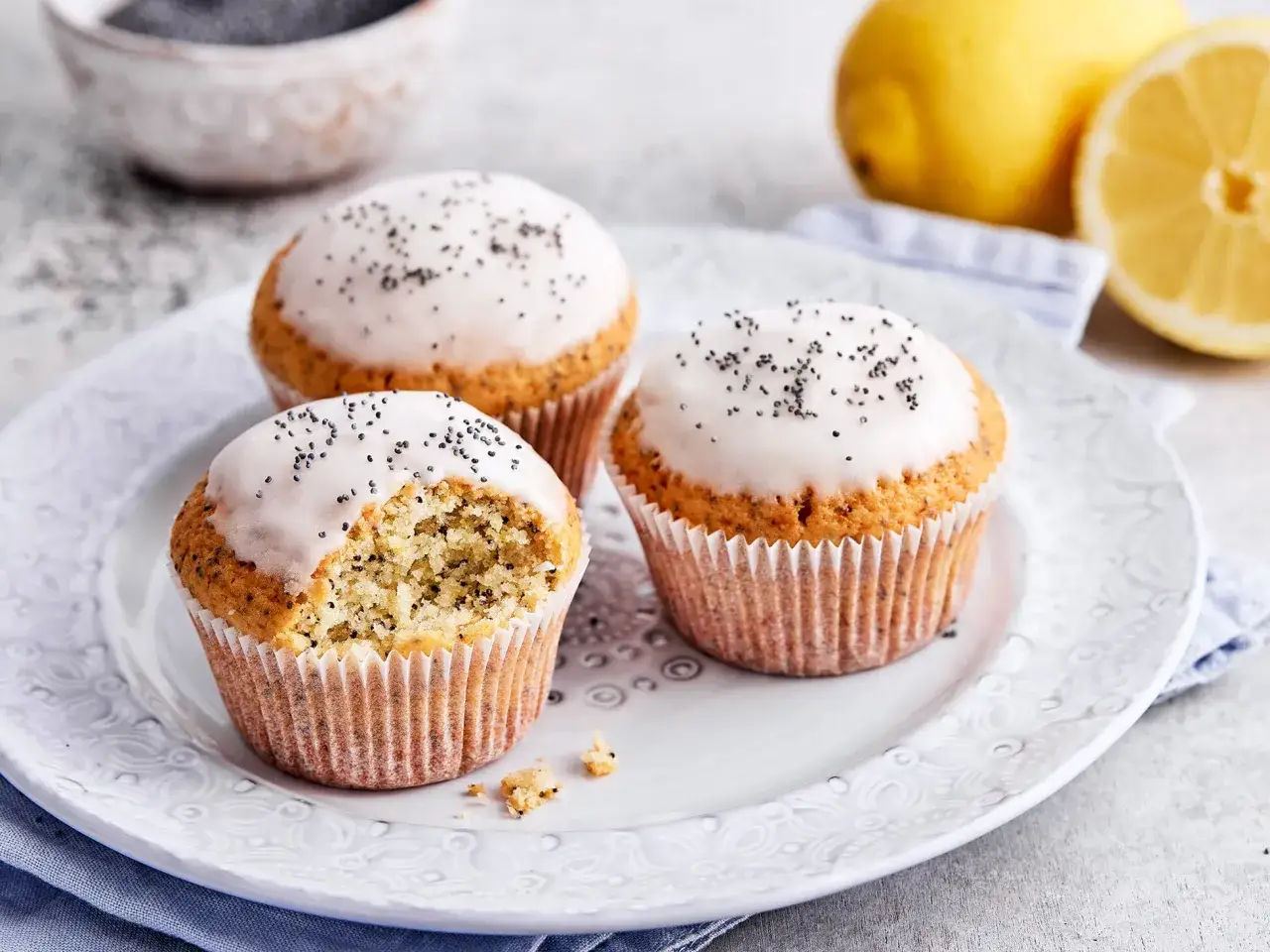 Drei zitronen mohn muffins mit wei&szlig;er Glasur und Mohn bestreut, einer davon angeknabbert. Frische Zitronen im Hintergrund.