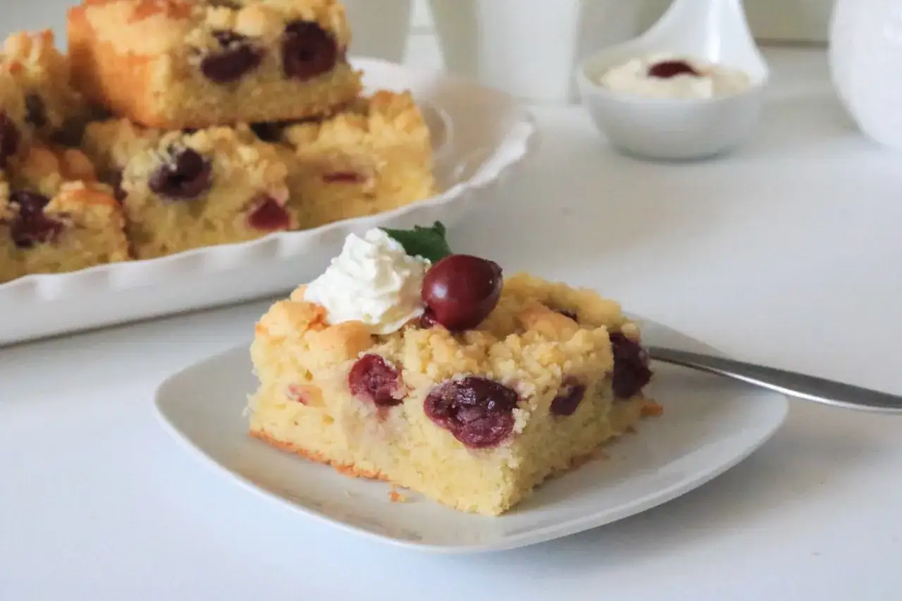 Ein Stück saftiger Kirschstreuselkuchen vom Blech, garniert mit Sahne und einer Kirsche.