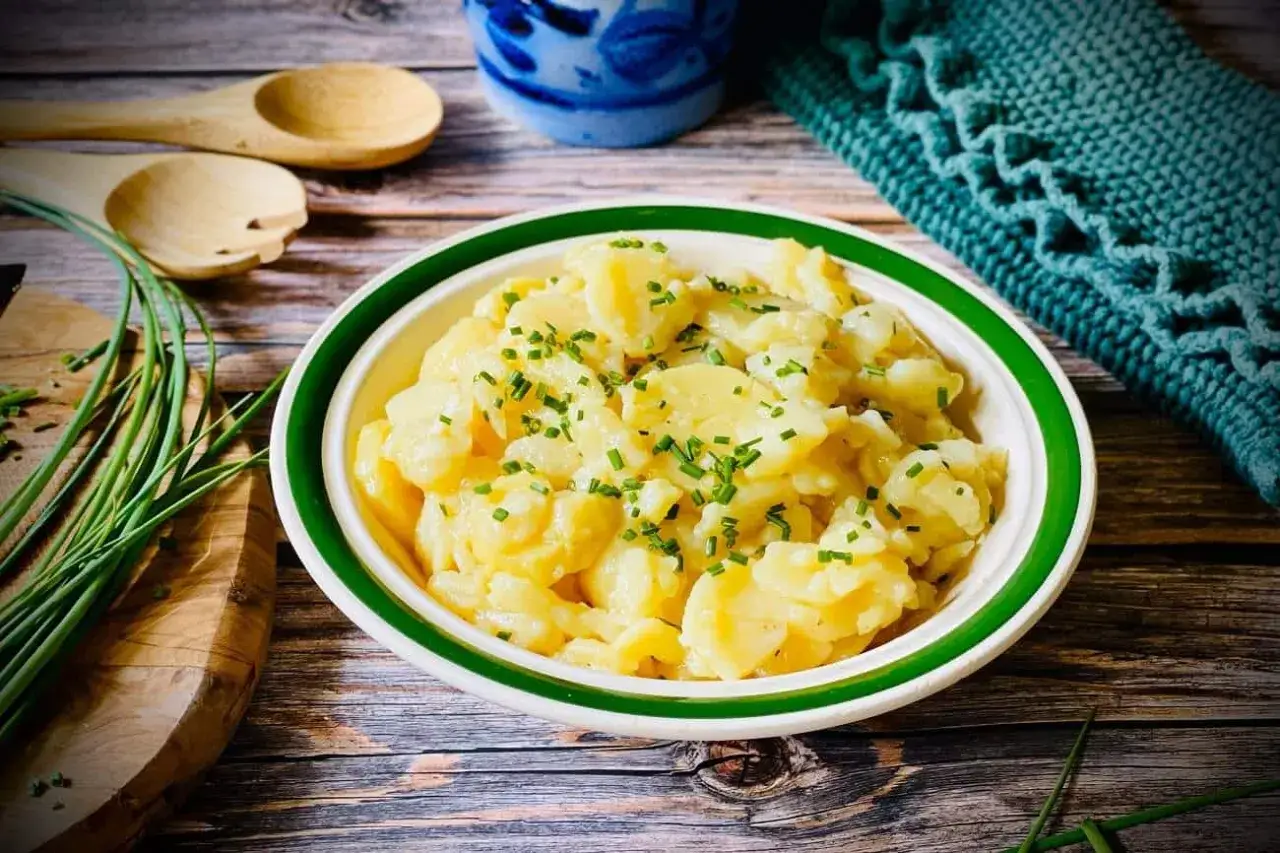 Sch&uuml;ssel mit Kartoffelsalat ohne Mayo, garniert mit Schnittlauch. Daneben liegen Holzkochl&ouml;ffel und ein gr&uuml;nes Tuch.