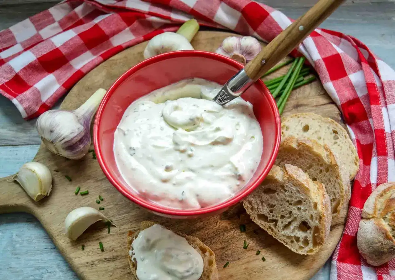 Cremiger Knoblauch Dip in roter Schüssel, daneben frisches Brot, Knoblauch und Schnittlauch. Perfekt zum Dippen!