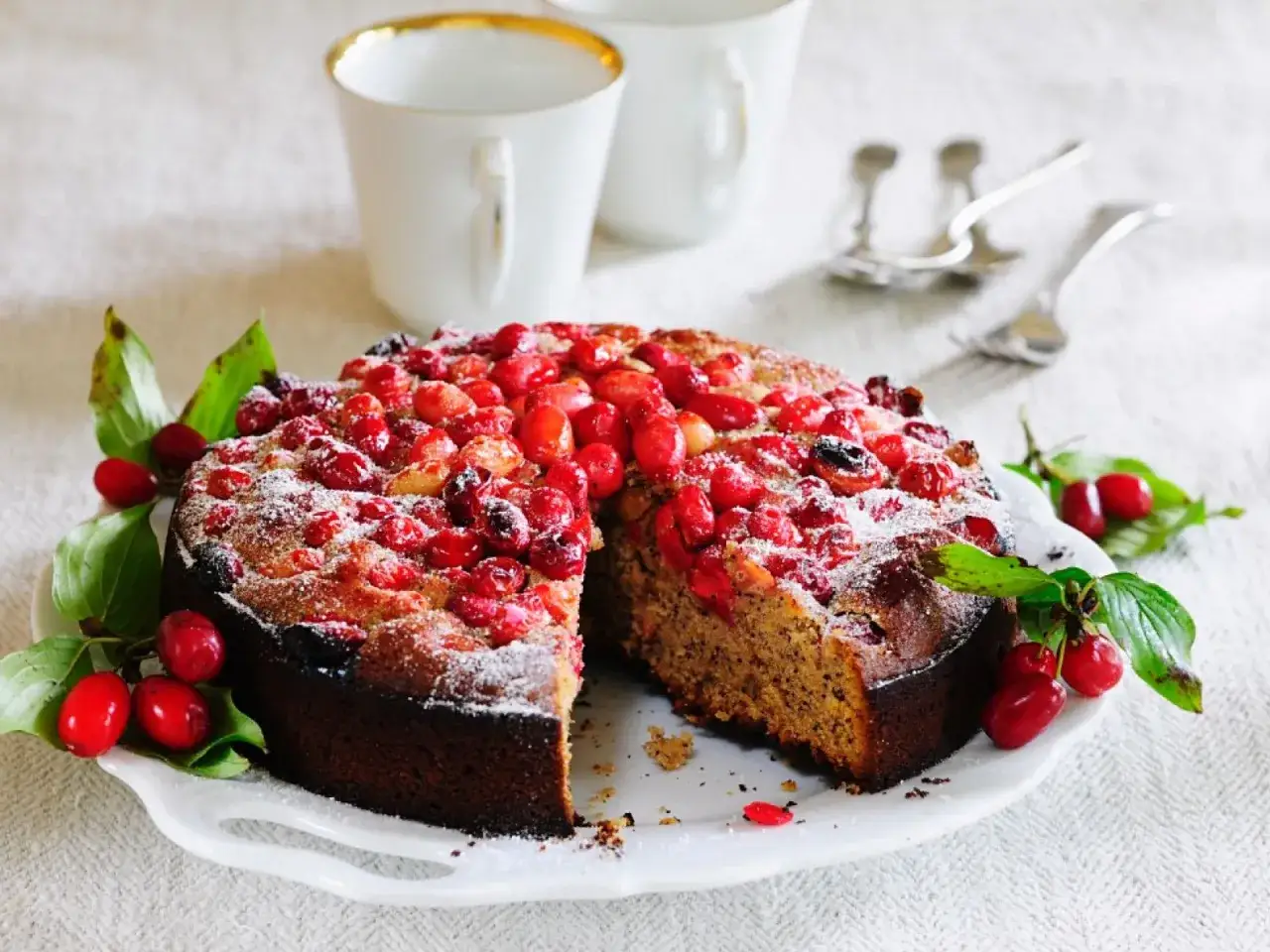 Kornelkirsche-Kuchen, ein k&ouml;stliches Rezept mit frischen Kornelkirschen, best&auml;ubt mit Puderzucker. Serviert mit zwei Tassen und Besteck.