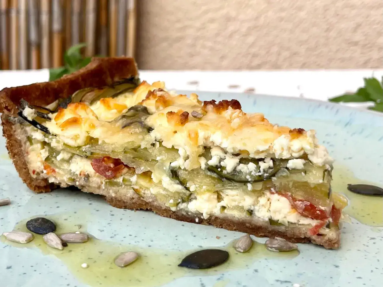 Ein St&uuml;ck herzhafte Zucchini Quiche mit K&auml;se und Tomaten auf einem blauen Teller, garniert mit Sonnenblumenkernen.