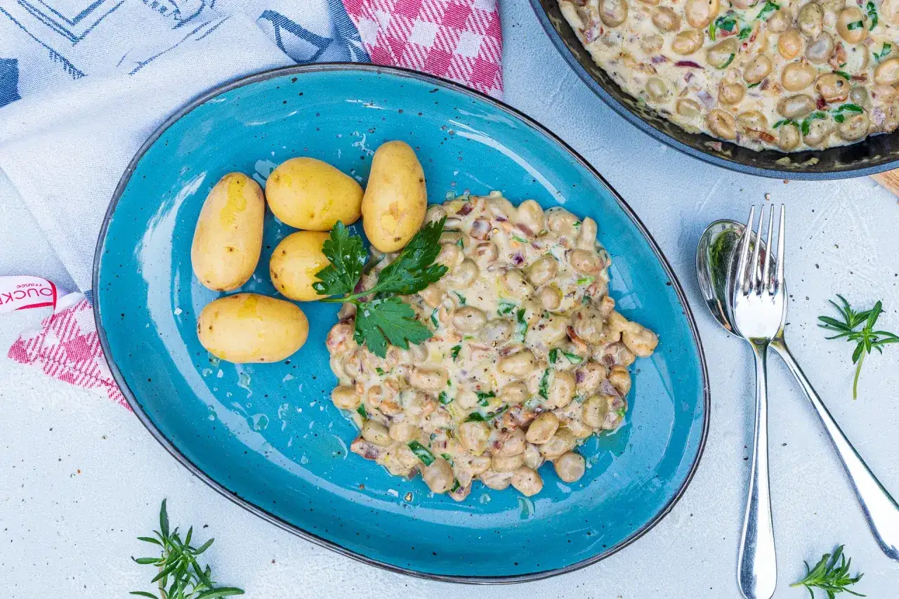 Cremiges dicke bohnen rezept mit kleinen Kartoffeln auf blauem Teller, garniert mit Petersilie. Daneben ein L&ouml;ffel und eine Gabel.