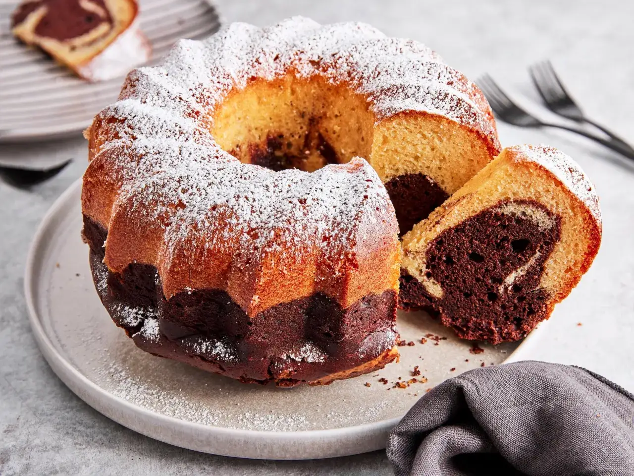 Ein saftiger Marmorkuchen Gugelhupf, bestäubt mit Puderzucker, wartet auf seine Genießer. Ein Stück ist bereits angeschnitten.