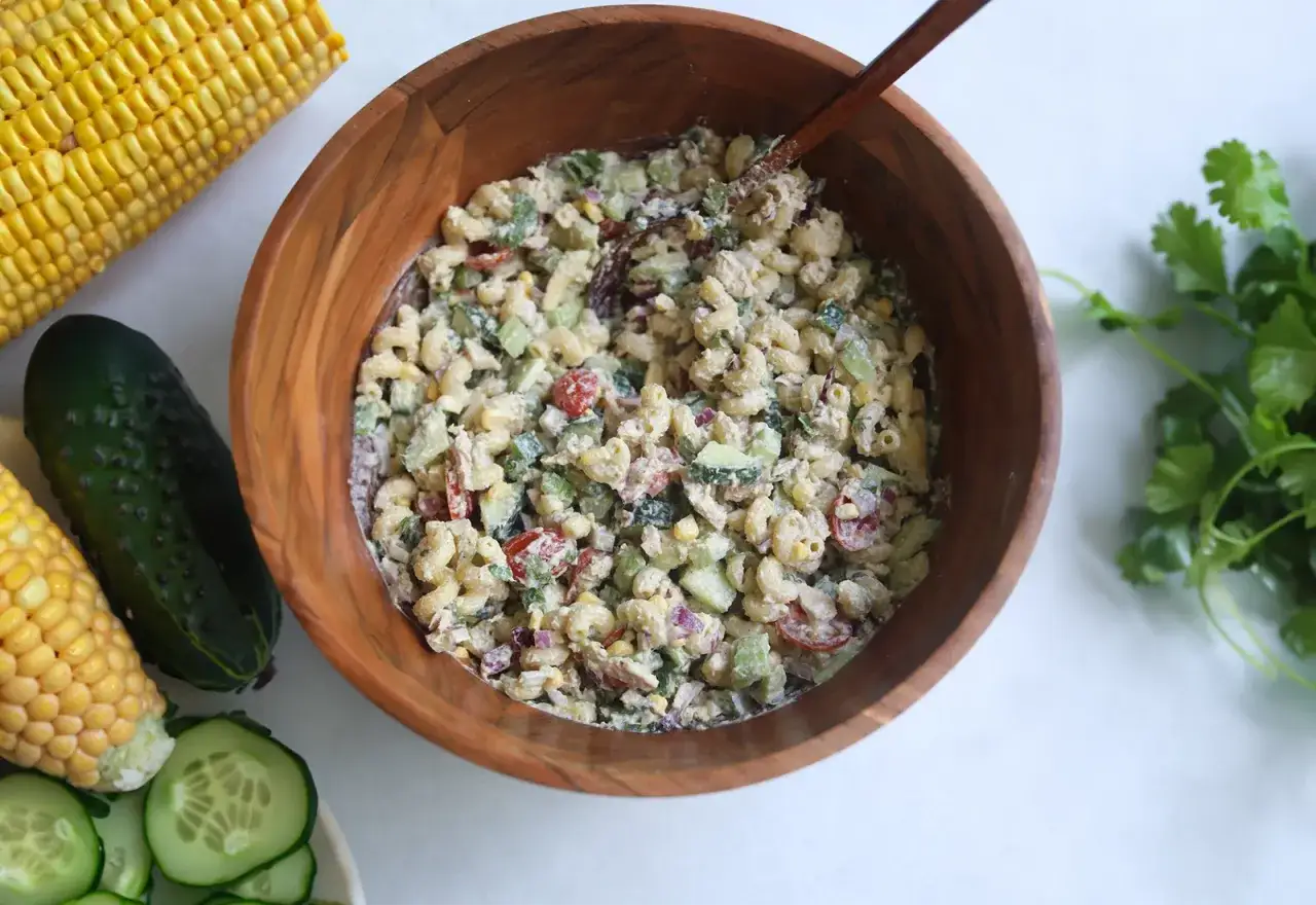 Herzhafter Thunfischsalat mit Nudeln, Gurken, Tomaten und Mais in einer Holzsch&uuml;ssel, daneben Maiskolben und frische Kr&auml;uter.