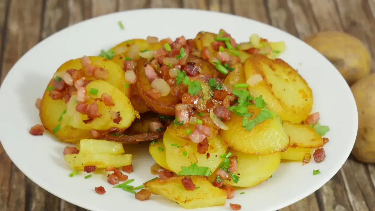 Goldbraune bratkartoffeln aus gekochten kartoffeln, garniert mit Speckw&uuml;rfeln und frischen Kr&auml;utern, auf einem wei&szlig;en Teller.