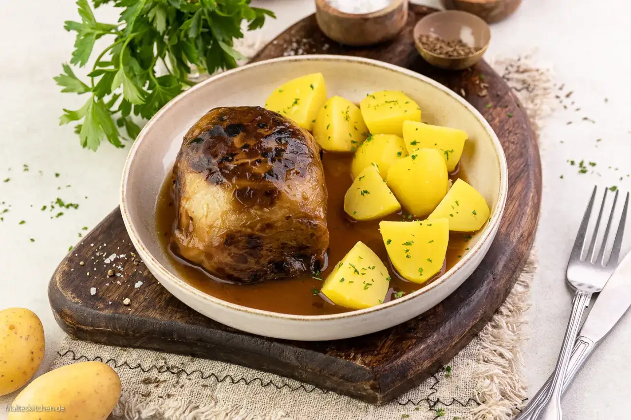 Herzhaftes Kohlrouladen Rezept: Ein goldbraun geschmorter Kohlrouladen-Strang mit Salzkartoffeln in einer kräftigen Sauce, garniert mit Petersilie.