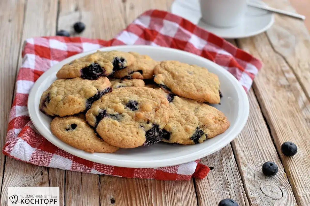 Saftige Blaubeer Cookies auf einem Teller, daneben frische Blaubeeren und eine rot-weiß karierte Serviette.
