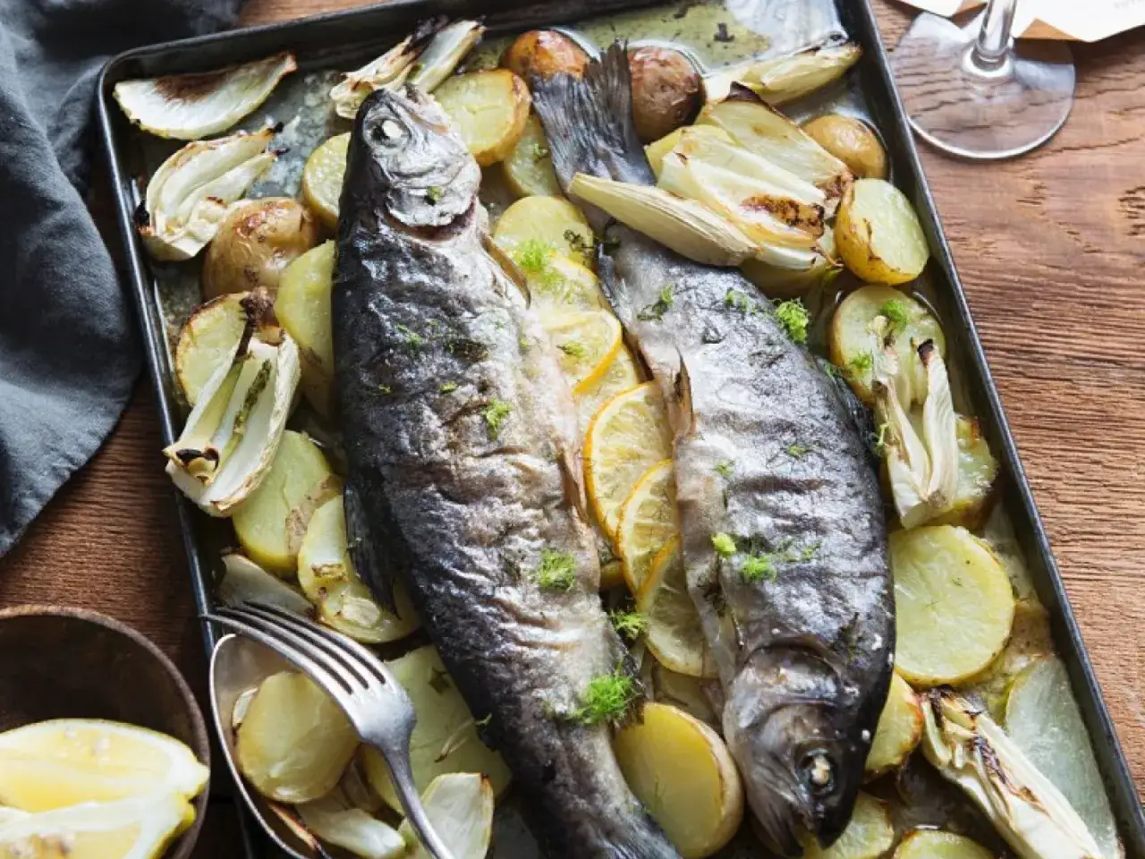 Zwei ganze Forellen im Backofen mit Kartoffeln, Fenchel und Zitronenscheiben auf einem Blech, garniert mit frischen Kräutern.